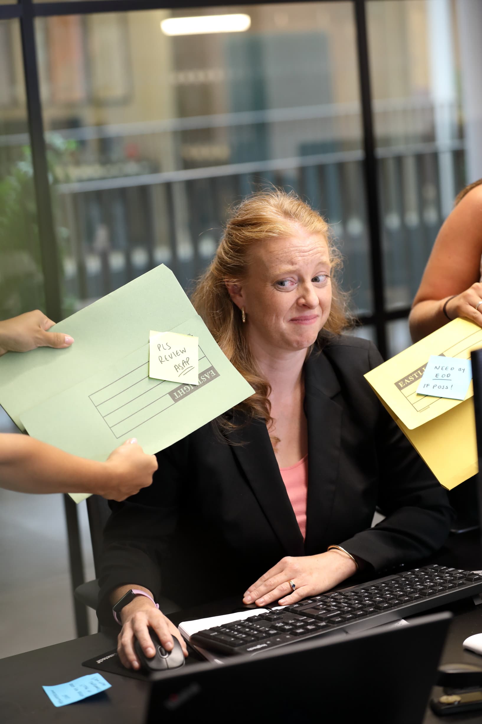 A woman at a desk looks overwhelmed as multiple people hand her folders labeled "Please review ASAP" and "Need by EOD if possible."