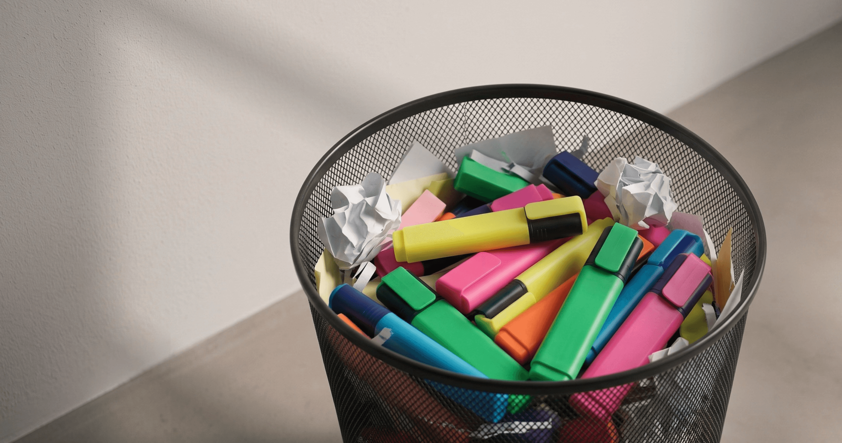 A wastebasket filled with colorful highlighters and crumpled paper on a light-colored surface.
