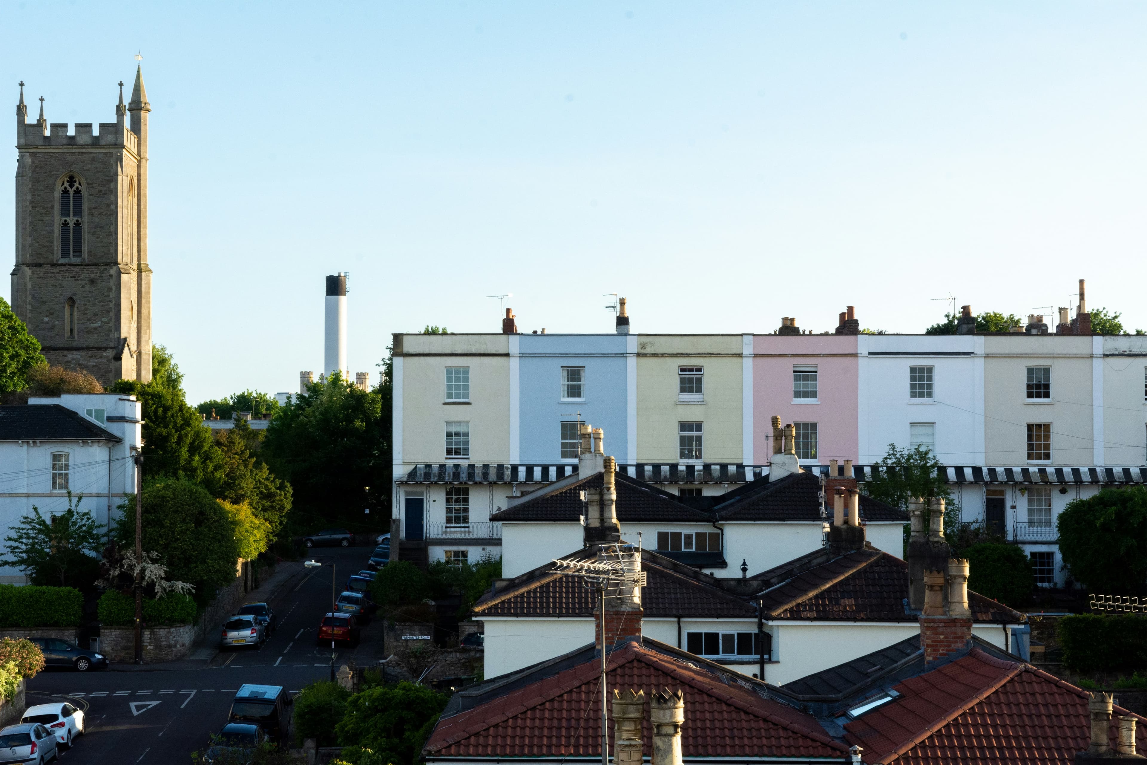 Street scene with pastel-colored row houses, a tall stone tower on the left, and a chimney in the background, under a clear blue sky.
