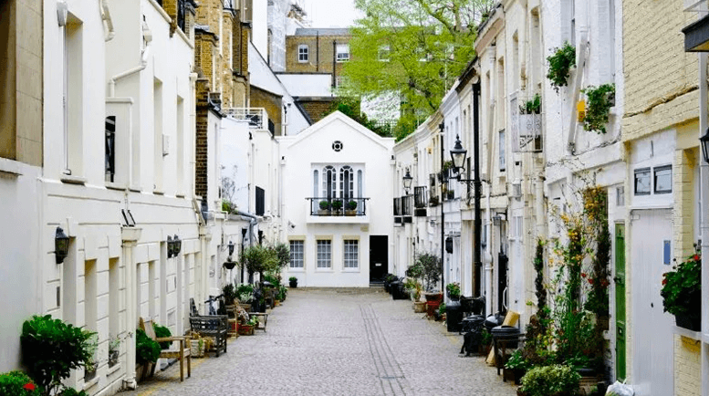 Narrow cobblestone alley lined with white townhouse-style buildings, adorned with potted plants and benches, leading to a central white house.